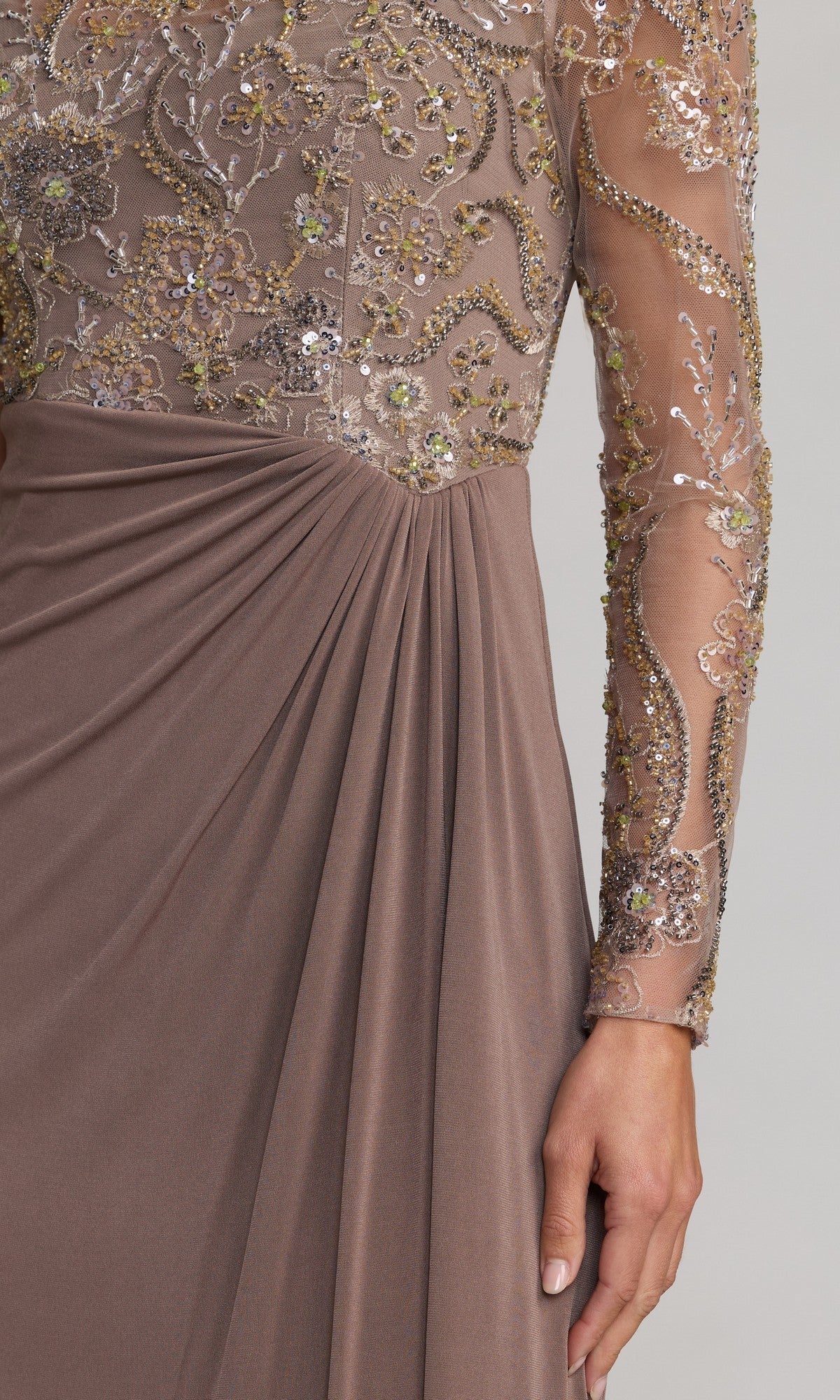 A close-up of a person wearing the Long Formal Dress: Mac Duggal 69121, a taupe evening gown with sheer long sleeves and an intricately sequin-embroidered bodice adorned with gold and silver floral patterns on the bodice and sleeves.